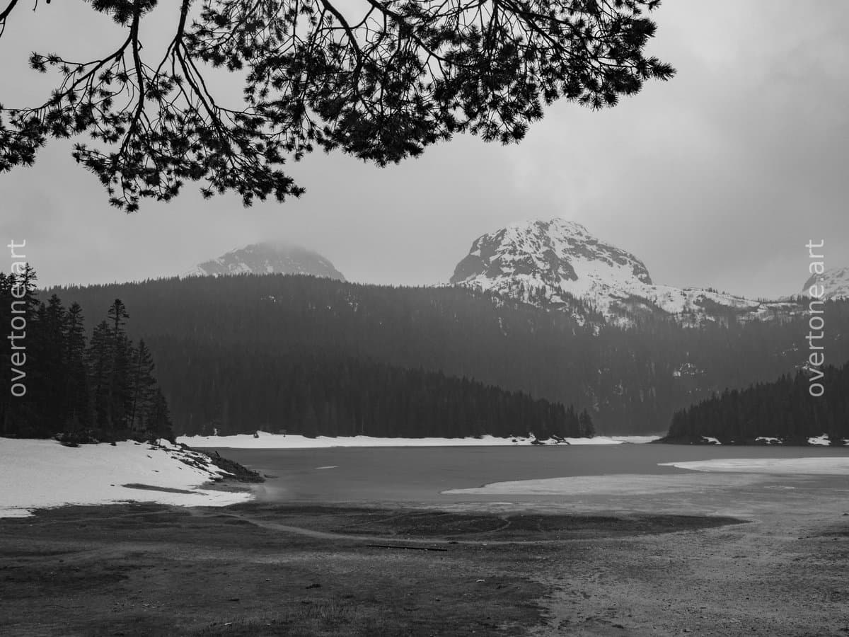 Black-and-white photograph of Black Lake (Crno Jezero) in Durmitor National Park, Montenegro, with a quiet shoreline, melting snow, evergreen forest, and snow-covered mountain peaks.