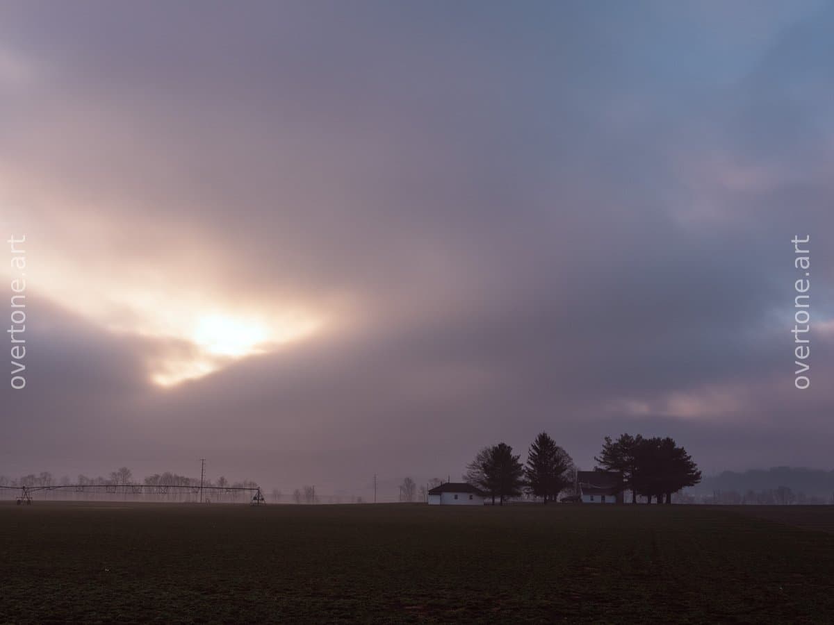 A low white farmhouse and a cluster of trees sit at the edge of a broad plowed field beneath a cloudy sky, with sunlight breaking through low on the horizon.